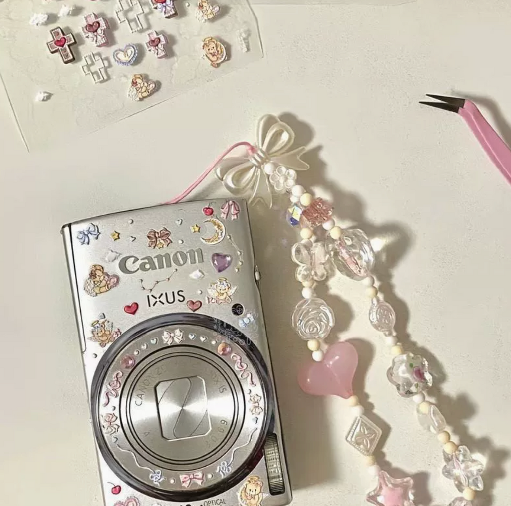 Coquette beaded camera charm strap styled beside a decorated compact camera